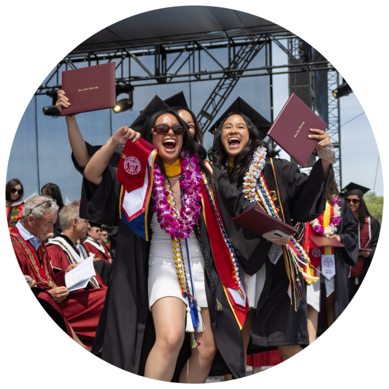Three students at graduation cheer on stage while holding their diploma up in the air.