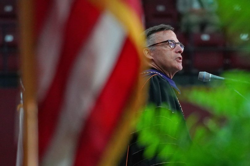 President Kevin OBrien speaks at his inauguration. His face is framed by a flag and plant. 
