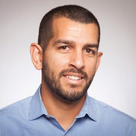 Andrew Ishak, smiling, wears a light blue shirt against a neutral background.