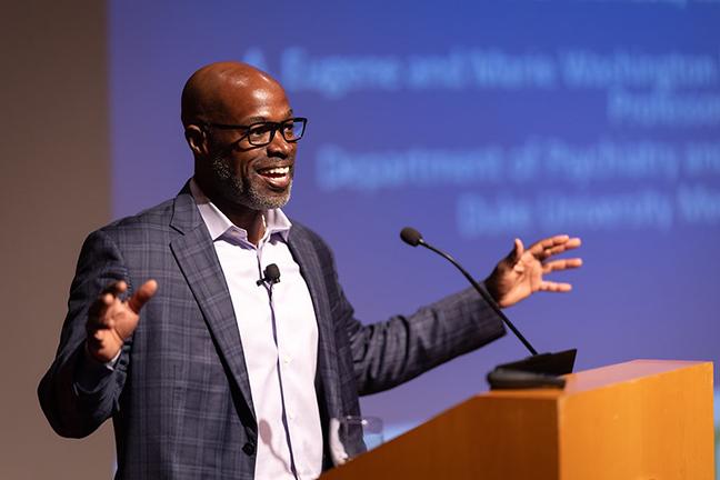 Kafui Dzirasa presenting a talk onstage at a podium in front of a blue screen with white text 