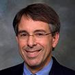 A person wearing glasses and a suit, smiling.