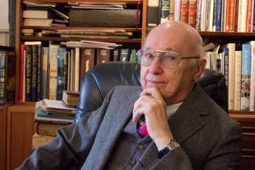 A man seated in an office with bookshelves behind him.