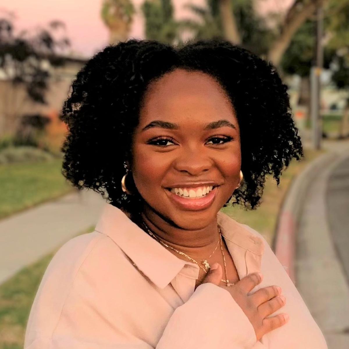 Person smiling warmly while walking outdoors at sunset, wearing a light-colored sweater.