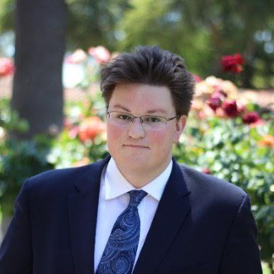 A person in a suit and tie stands in front of flowers.