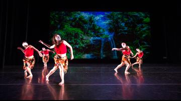 5 female dancers in red costumes performing on stage with tropical backdrop