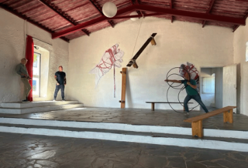 2 men stand on the left and 1 man stands on the left of a large room with cream walls, a wooden roof and grey tiled floor.  Theres a bench on the right and modern art on the far wall.