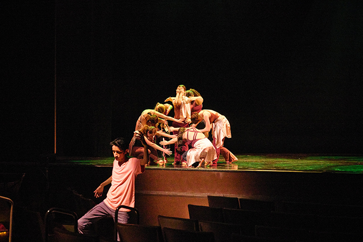 A group of female dancers huddled together onstage with a male dancer sliding off the stage into the audience