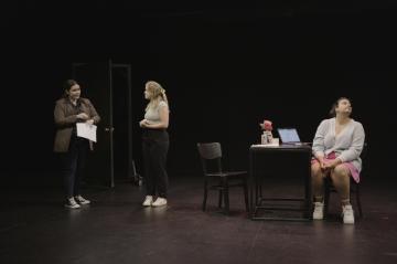 Ina black box theatre, one woman entered through a door to meet a second woman whos standing and a third whos seated in the bcakground