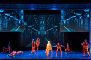 Stage performance of Legally Blonde with actors in pink-orange outfits against a blue backdrop.