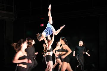6 female dancers in black lifting overhead a female dancer in a pale blue dress