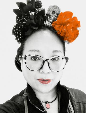 Headshot of female wearing glasses with large flowers and skull in her hair. The photograph is black and white except for orange on her lips, necklace, and one of the flowers.