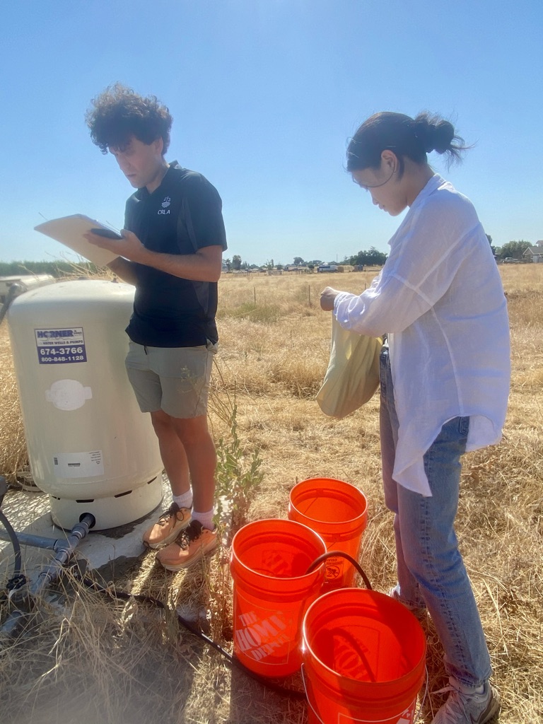 Two people conducting a water test in a dry field, standing next to a large white tank and orange buckets.