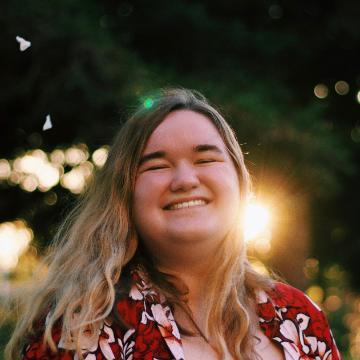 A woman smiling outdoors with sunlight and trees in the background.