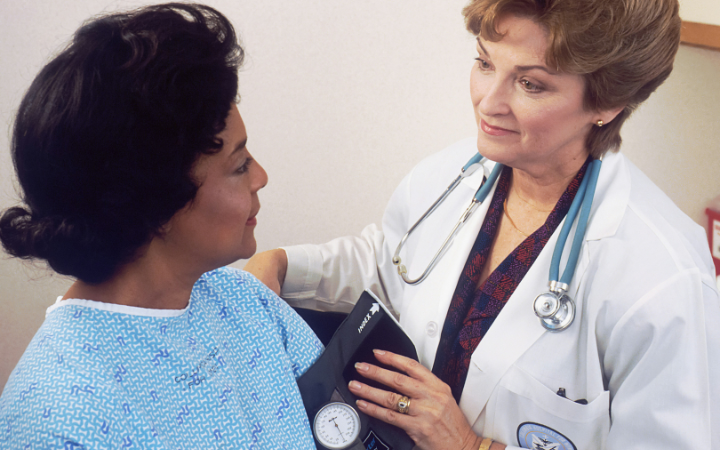 A woman with a stethoscope and wearing a medical coat talking to another woman wearing a patients gown.