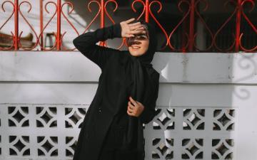A woman wearing a hijab standing against a white wall with red iron fencing uses her hand to block sunlight from her eyes. Image by Amrullah Ab via Pixabay.