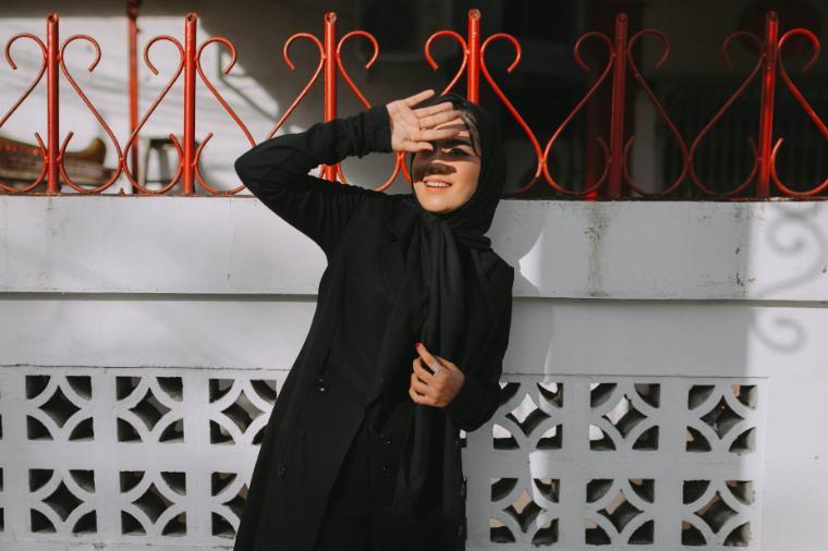 A woman wearing a hijab standing against a white wall with red iron fencing uses her hand to block sunlight from her eyes. 