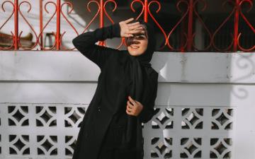 A woman wearing a hijab standing against a white wall with red iron fencing uses her hand to block sunlight from her eyes. Image by Amrullah Ab via Pixabay.