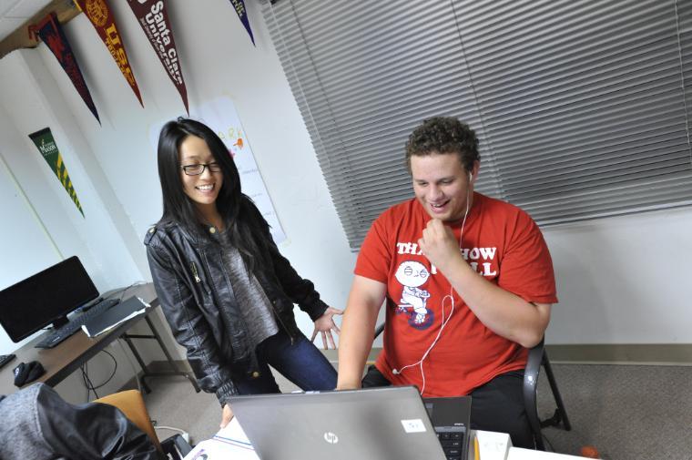 A person in a red shirt sits at a desk working on a computer while another person in black stands nearby.