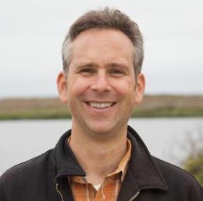 A person standing near a body of water, smiling.