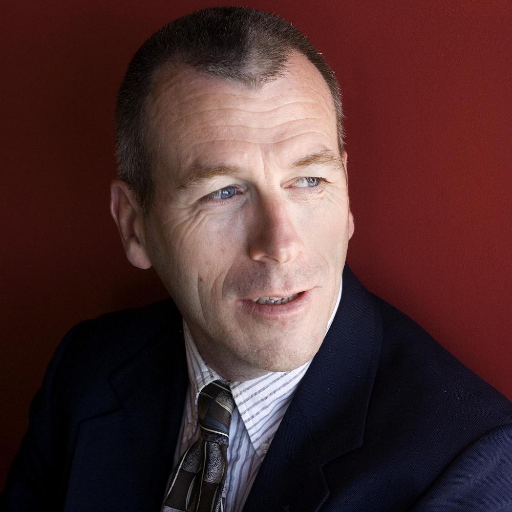A person in a suit looking to the side against a red backdrop.