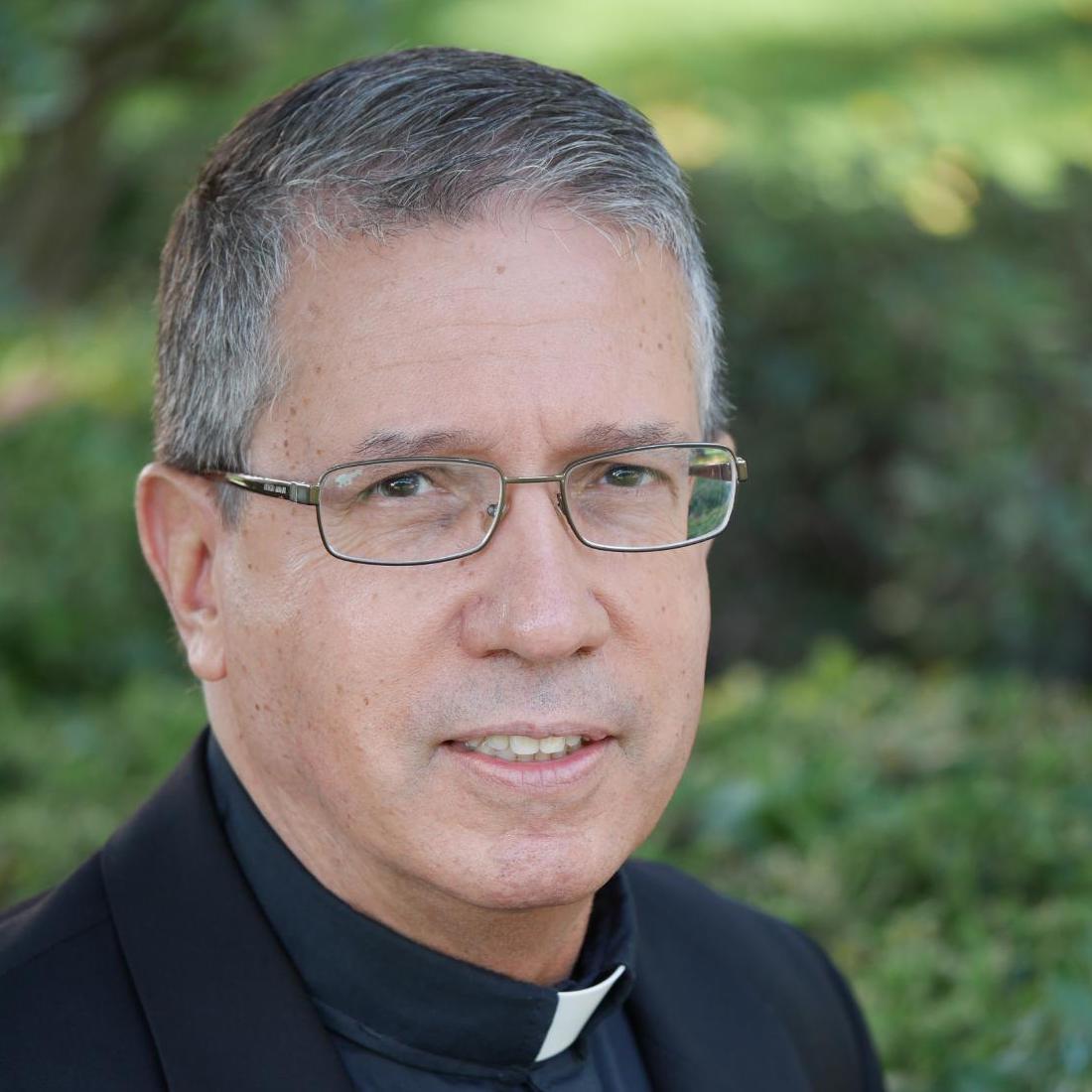A man in clerical clothing, outdoors, with greenery in the background. 