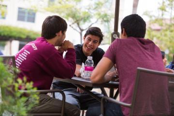 Three individuals sitting outdoors, talking, and enjoying beverages at a table.