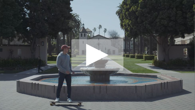 Man with a skateboard standing by a fountain, white play button overlay.