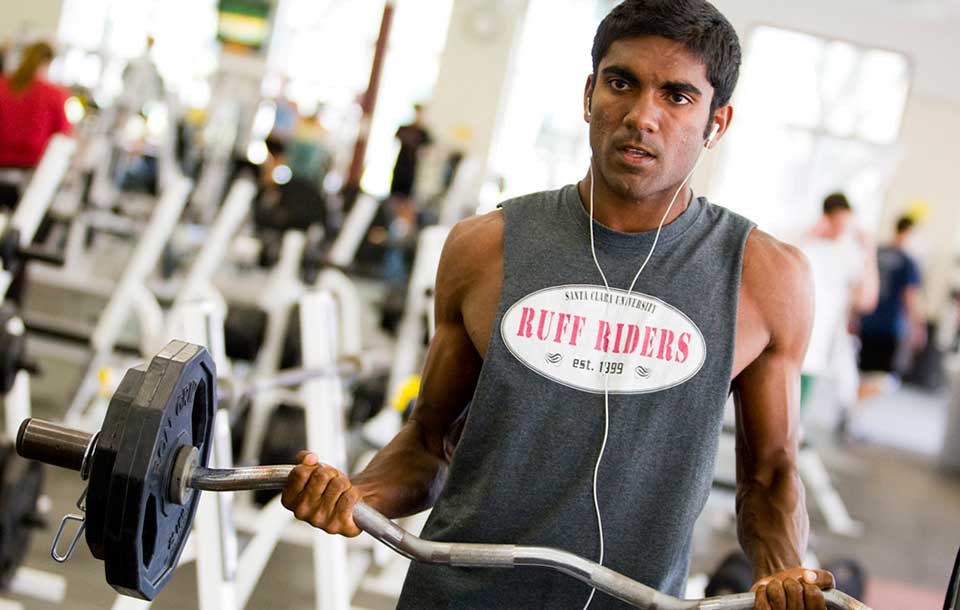 Man lifting a barbell in a gym wearing a sleeveless shirt reading NO SMOG.
