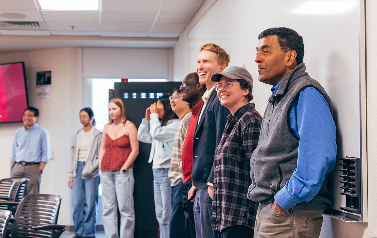 A group of faculty and students standing up against a white board on the right side of the frame, looking off frame to the left with smiling expressions.