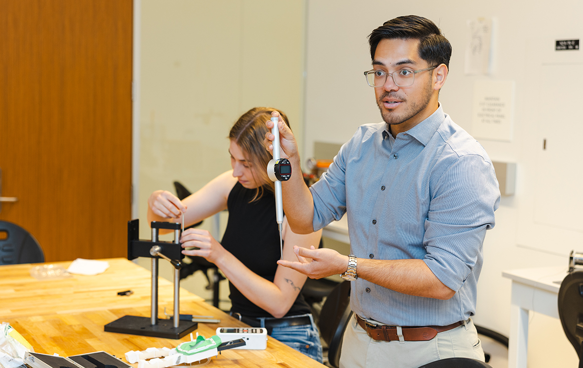 Michael Abbott holds metal tube instrument over lab table with student working at table in background.