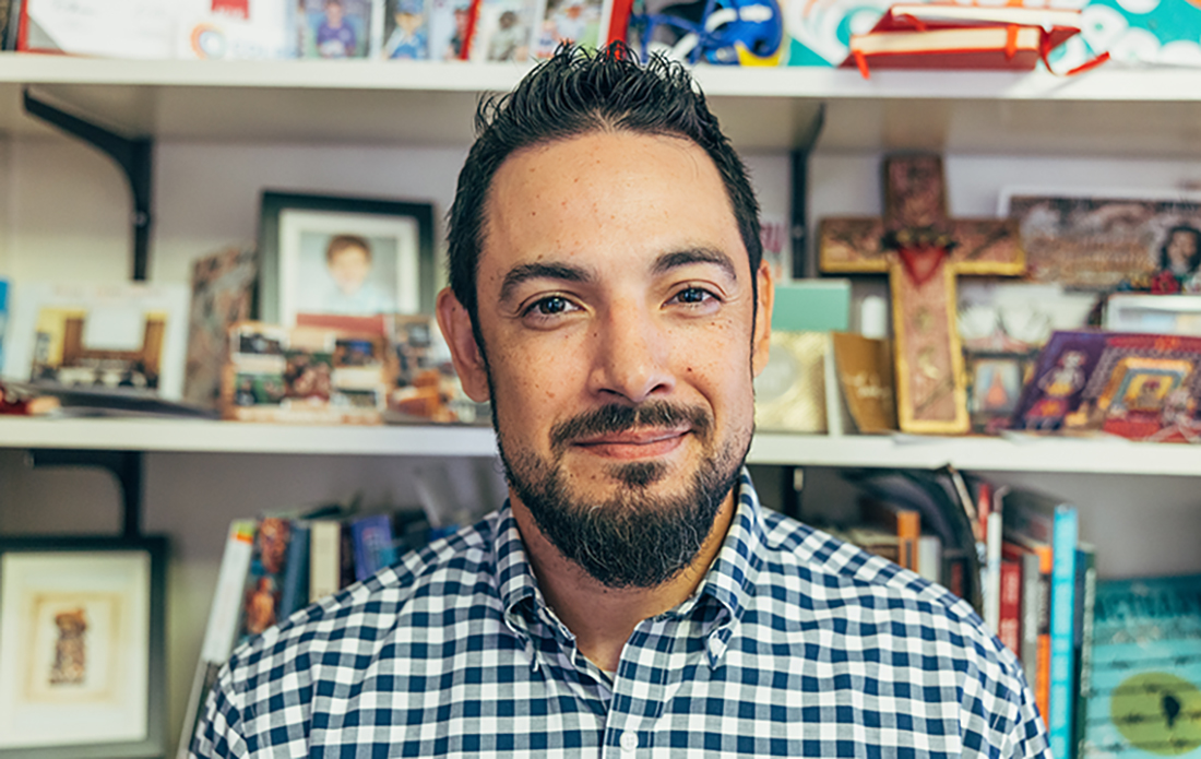 Man with brown hair and beard wearing gingam pattern button up shirt sits in office with busy shelves in the background. On the shelf, a cross is prominently displayed.
