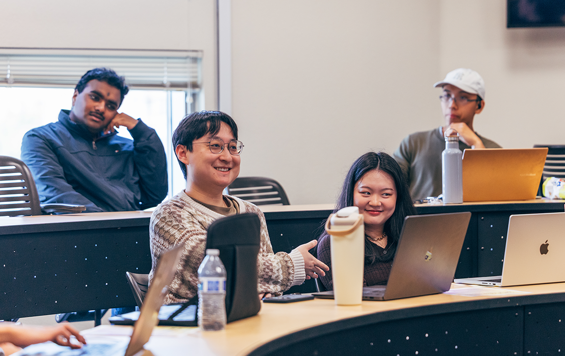Several students sit with laptops at long concentric tables in a lecture hall, two smiling.