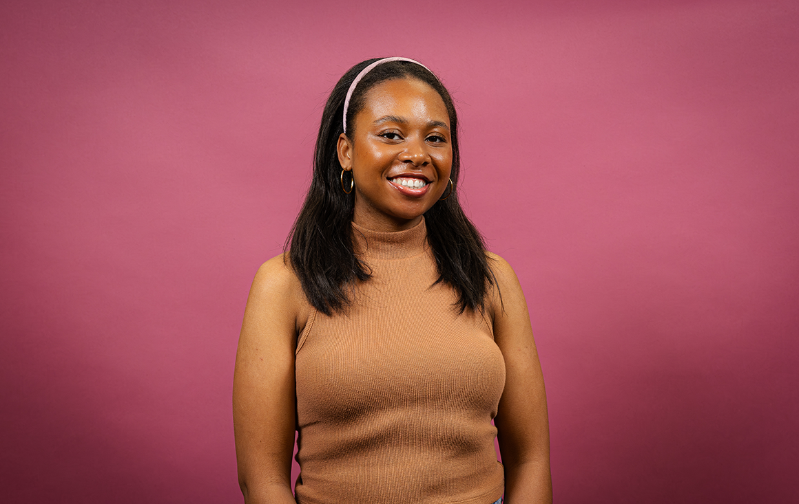 A young Black woman in a peach, sleeveless blouse stands in front of a pink backdrop.