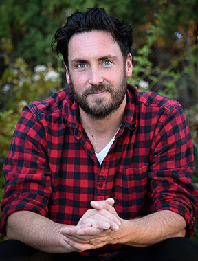 Author JP Lacrampe in a red flannel shirt in front of plants and trees.