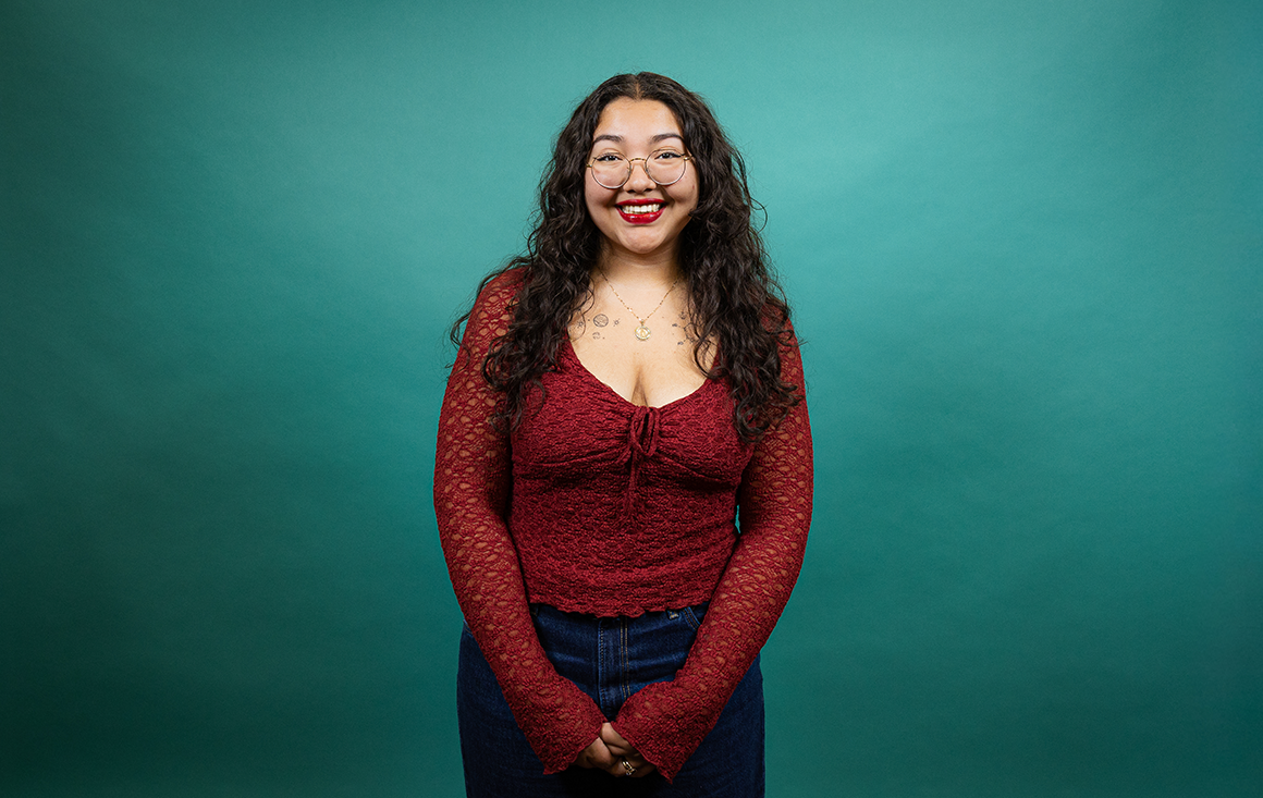 A young Latina woman with long, curly black hair wearing a red blouse, denim jeans, and glasses stands in front of a teal backdrop.