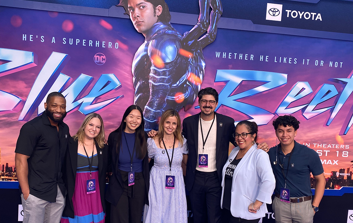 A group of people pose shoulder-to-shoulder in front of a Blue Beetle promotional poster.