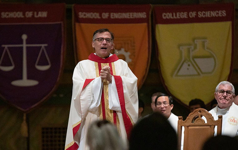 Fr. Kevin OBrien speaks in the Mission Church with academic flags hanging in the background