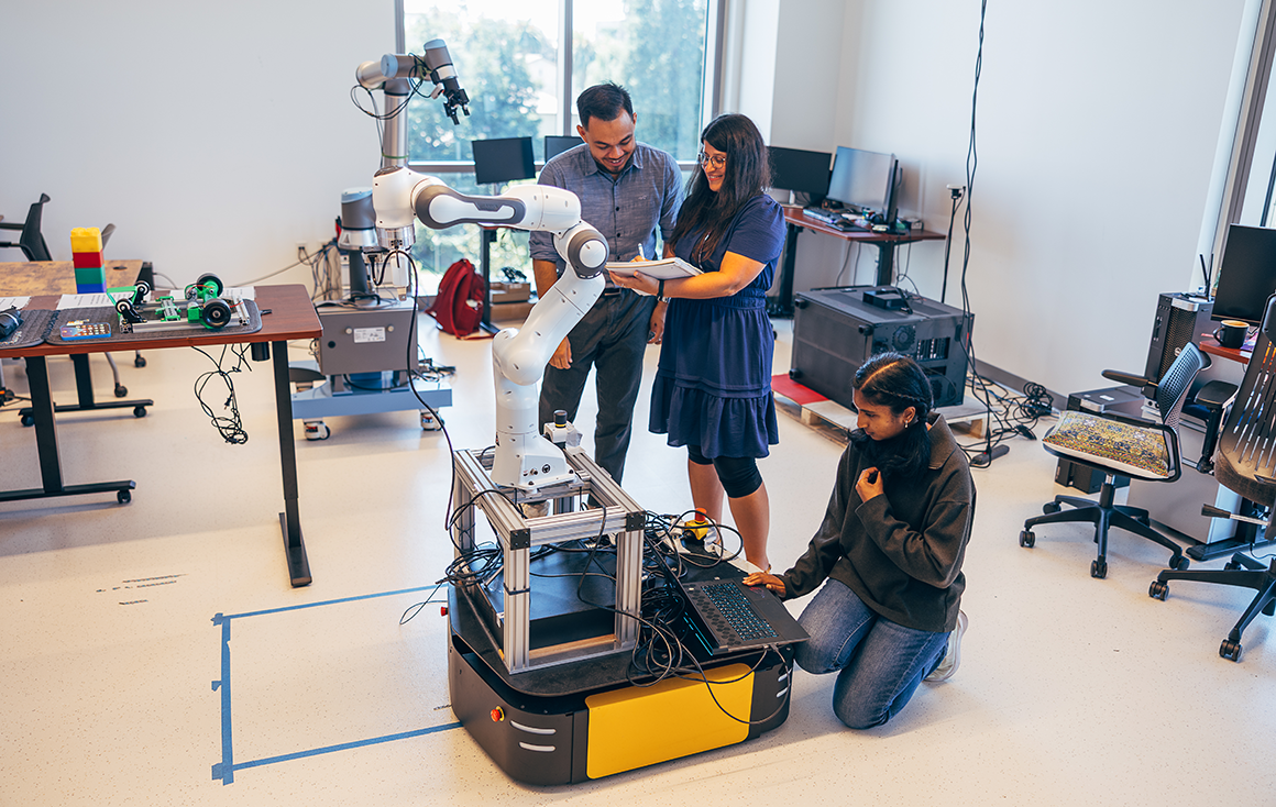 A teacher holding an iPad stands with two students in a robotics lab with a large robotic arm on a mobile platform.