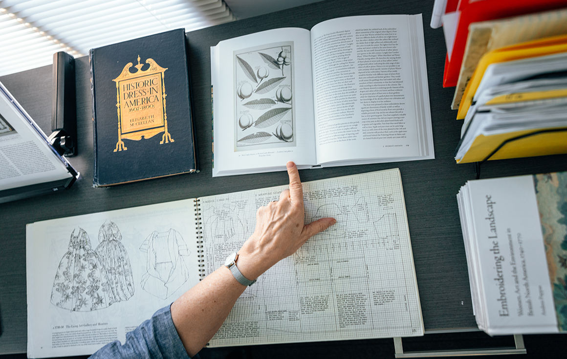 A womans hand point to a stone fruit illustration in an open book on a desk.