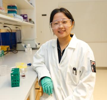 In the chemistry lab, An analyzes a nanoparticle sample. A female student wearing a lab coat and goggles at a counter in a chemistry lab.
