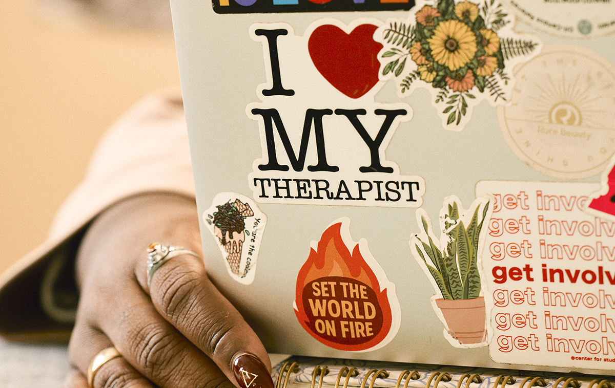 A womans hand peeking around the edge of a laptop with a variety of stickers including I heart my therapist and set the world on fire.