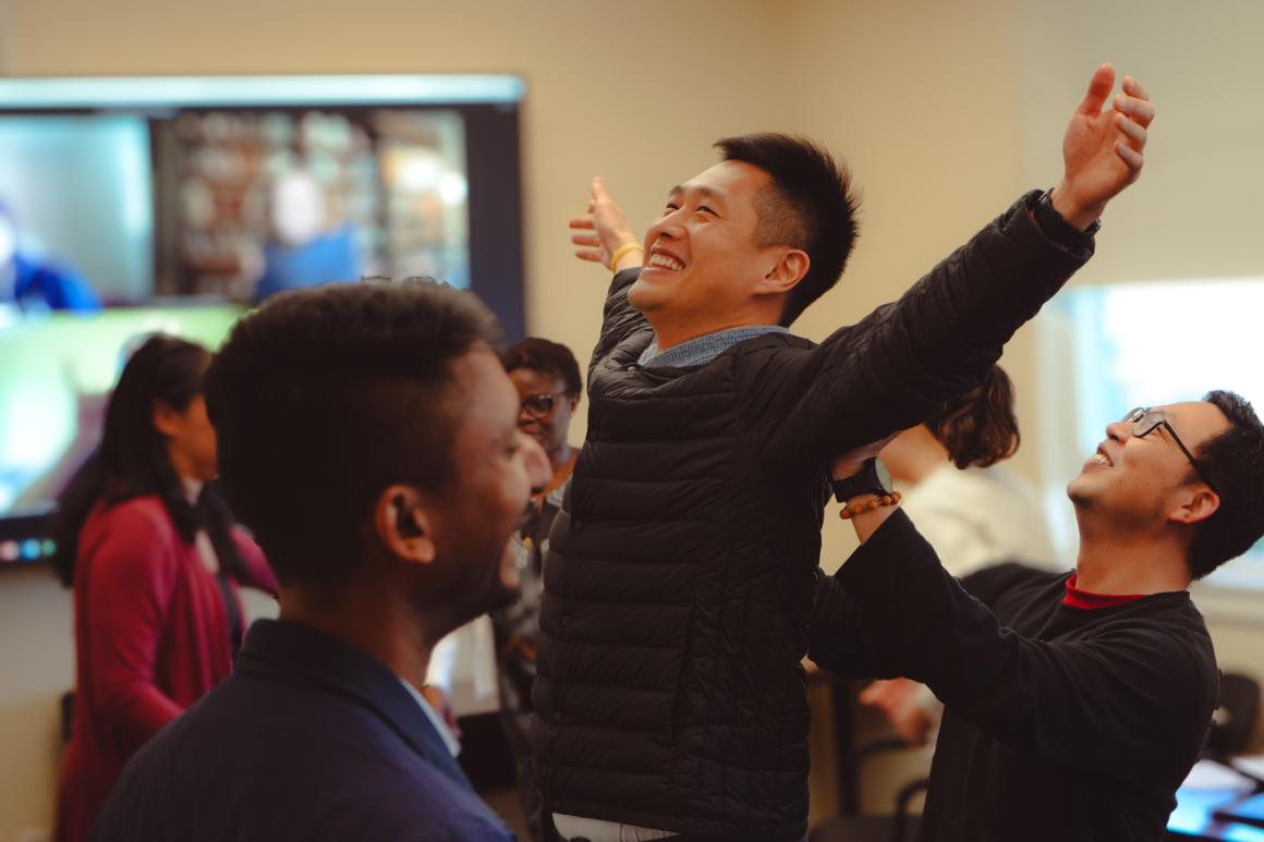A student smiling in a crowded classroom. Their arms are stretched wide and they appear to be doing a trust exercise with another student behind them.
