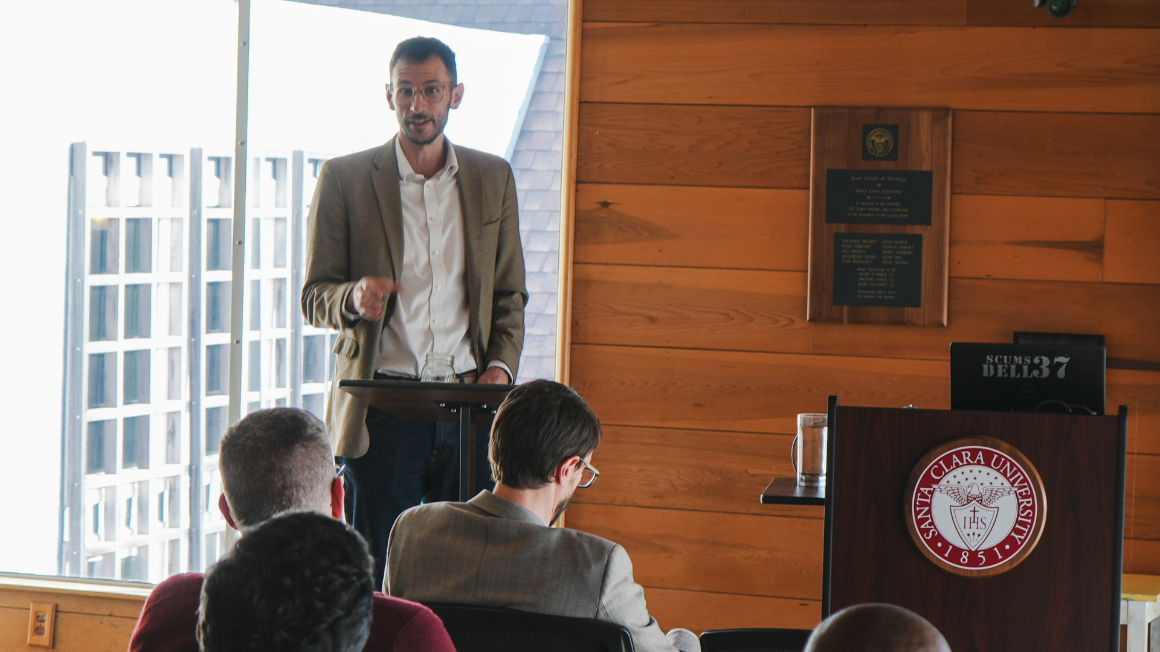 A man giving a lecture. There are people seated in the foreground. The background has a large window and wood paneled wall.