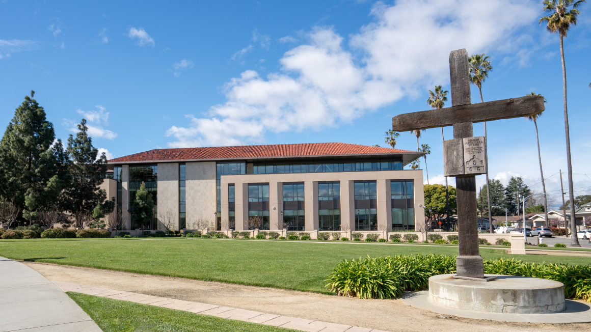 A wooden cross with an academic building in the background. There is green grass and a blue sky with clouds.