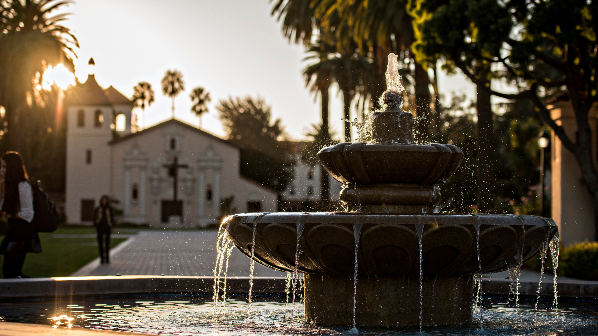 A three-tiered fountain is in the foreground. There is a Spanish Mission style church in the background. Palm trees surround the area.