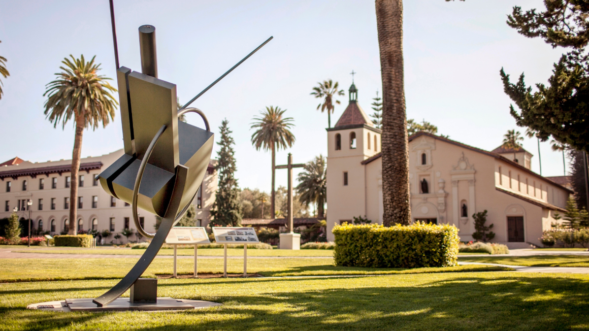 A photo of a college campus. There is a blue sky and green grass. The foreground has an abstract sculpture, and the background has a Spanish style mission church.