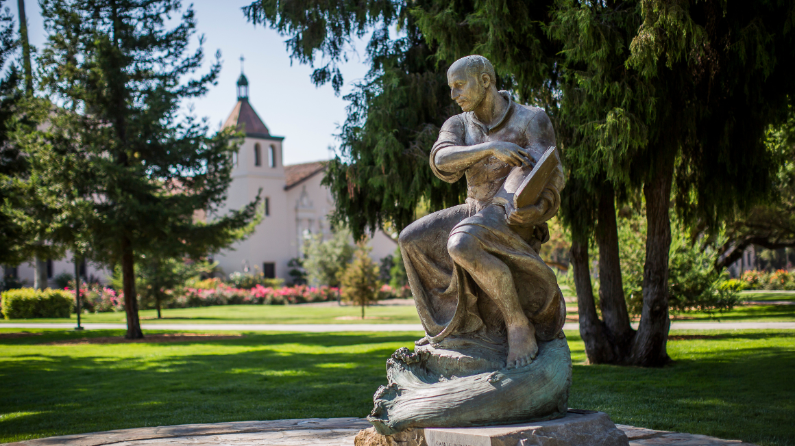 A bronze statue of St. Ignatious in a contemplation pose. There are evergreen trees and a Spanish mission style church in the background. A fountain is at the base of the statue.