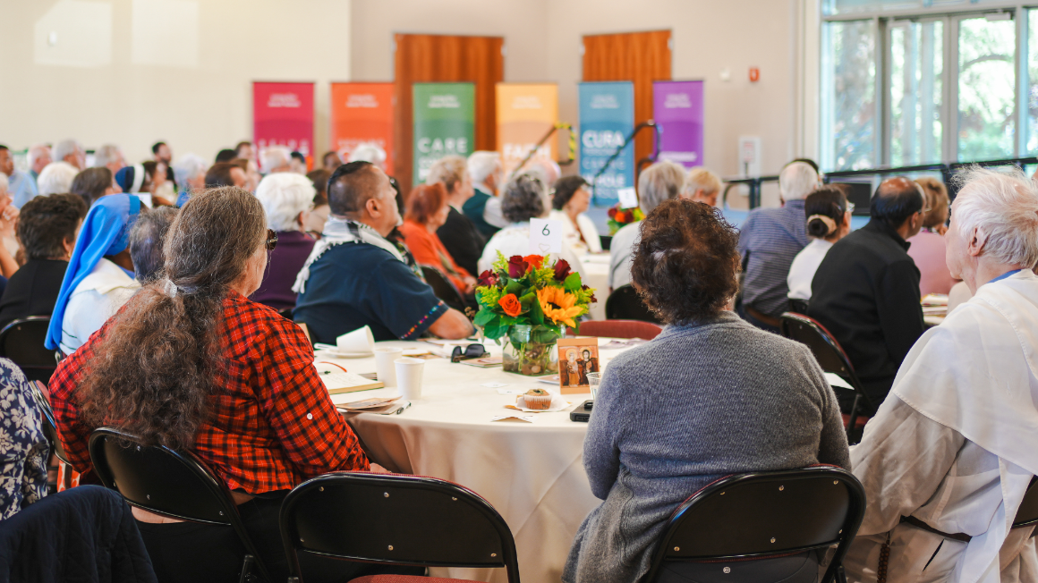 People sitting at round tables in a large conference room with colorful banners.