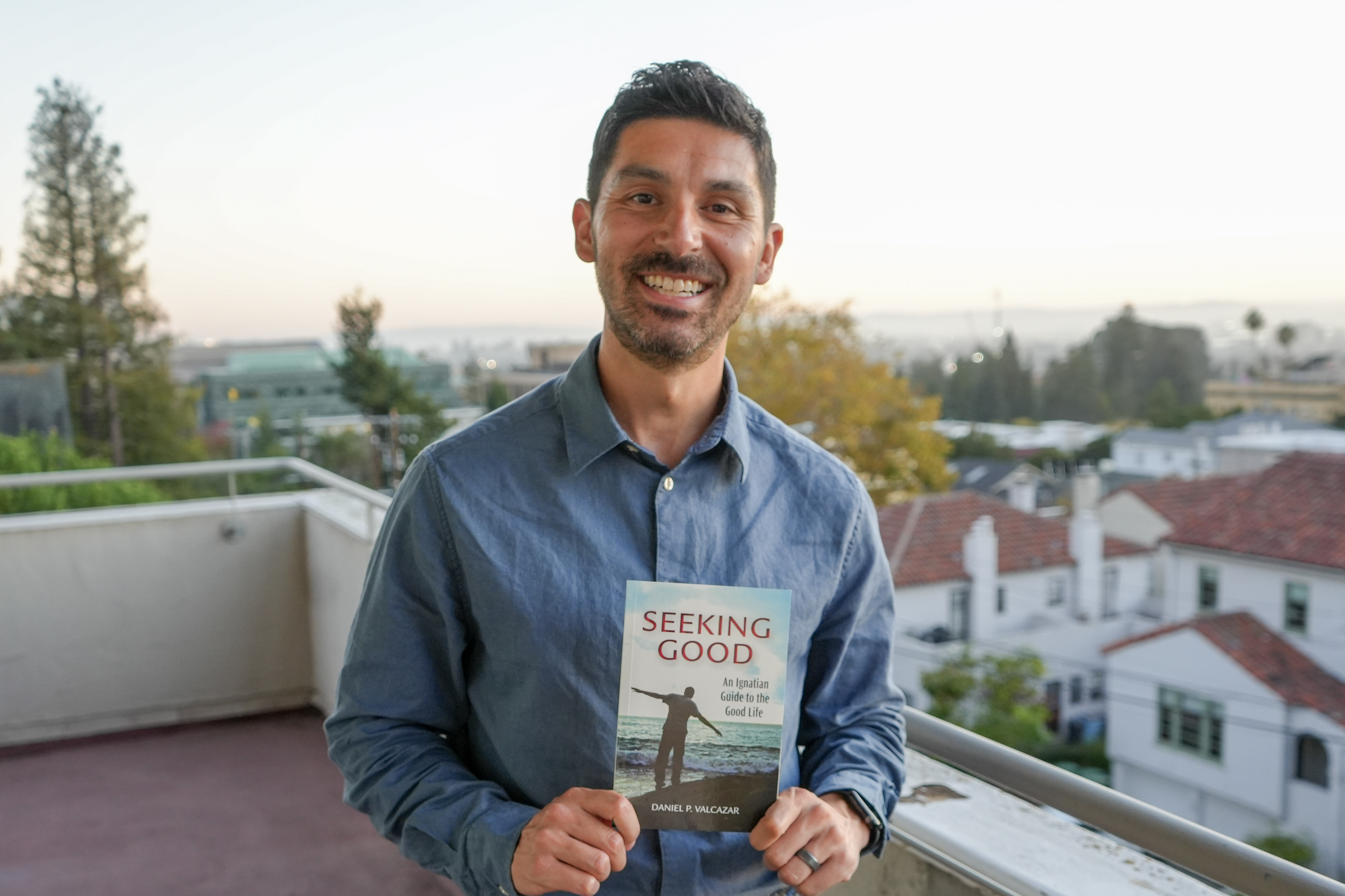 A man stands on a balcony holding a book and smiling. The book is titled Seeking Good: An Ignatian Guide to the Good Life. The author is listed as Daniel P. Valcazar.