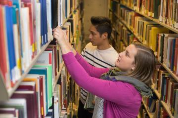 two students stand next to each other searching for a book on a bookshelf in the library